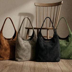 May include: Four suede shoulder bags in shades of brown, gray, black, and green stand in front of a wooden chair. The bags have a crescent shape with long, thin handles. The bags are displayed on a wooden floor.