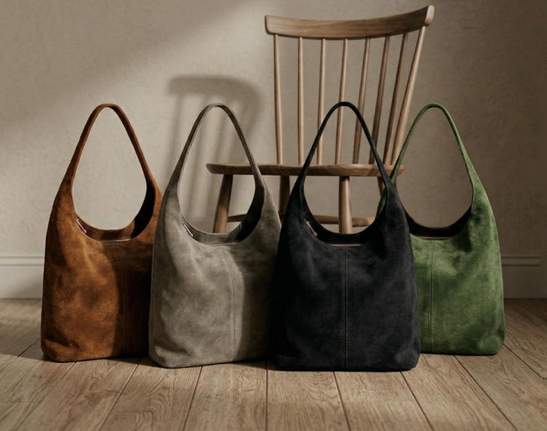 May include: Four suede shoulder bags in shades of brown, gray, black, and green stand in front of a wooden chair. The bags have a crescent shape with long, thin handles. The bags are displayed on a wooden floor.