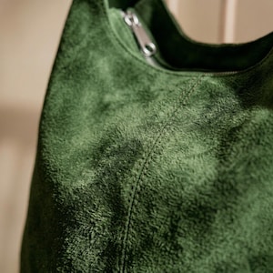 May include: Close-up of a green suede shoulder bag. The bag has a zipper closure and a stitched seam detail. The suede material has a textured, plush appearance. The bag is a solid, deep green color.
