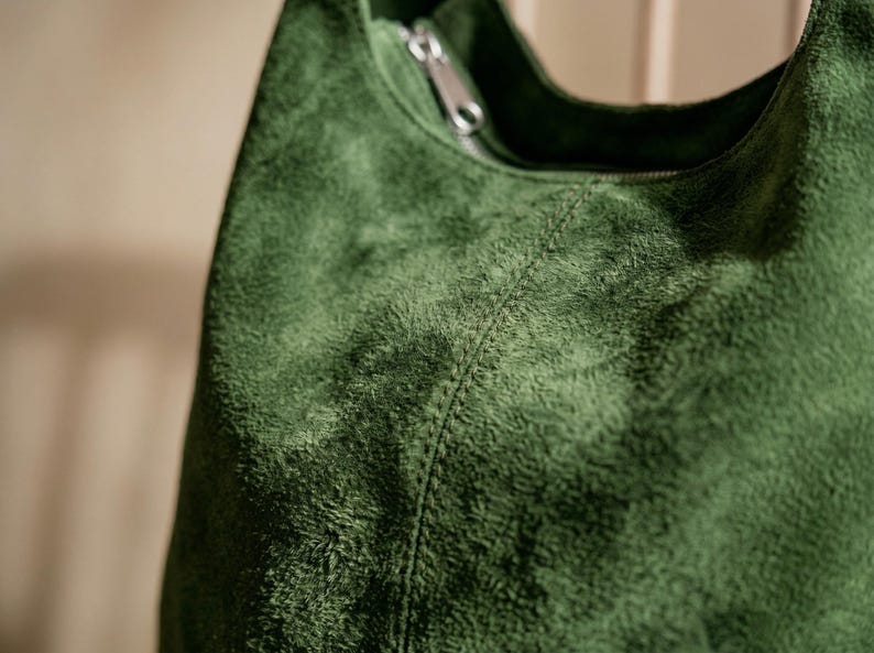 May include: Close-up of a green suede shoulder bag. The bag has a zipper closure and a stitched seam detail. The suede material has a textured, plush appearance. The bag is a solid, deep green color.