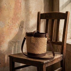 May include: A bucket bag featuring a textured beige woven body with dark brown leather trim at the top and bottom. The bag includes a dark brown leather handle, a matching adjustable shoulder strap, and a gold-toned metal clasp closure.