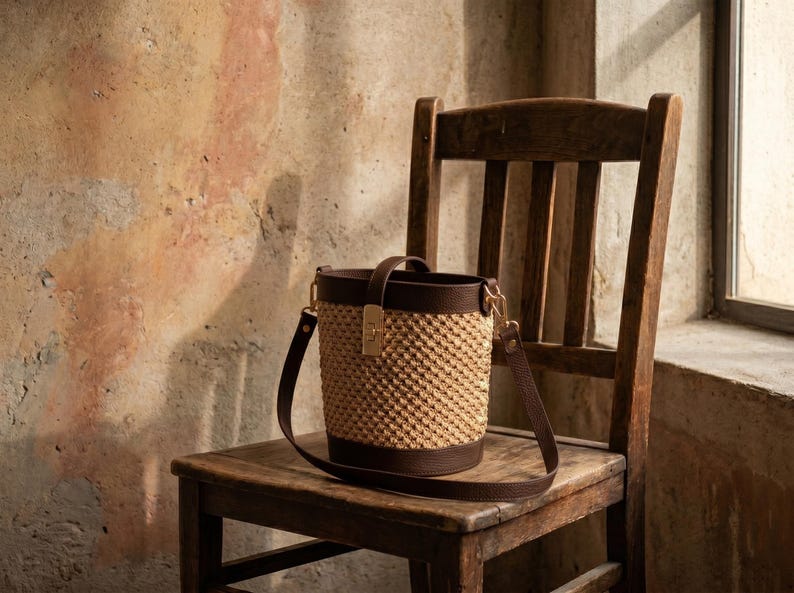 May include: A bucket bag featuring a textured beige woven body with dark brown leather trim at the top and bottom. The bag includes a dark brown leather handle, a matching adjustable shoulder strap, and a gold-toned metal clasp closure.