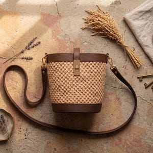May include: A bucket bag featuring a woven tan straw body with dark brown leather trim at the top and base. The bag includes a gold-tone metal turn-lock closure on a vertical leather strap and a long, adjustable dark brown leather shoulder strap.