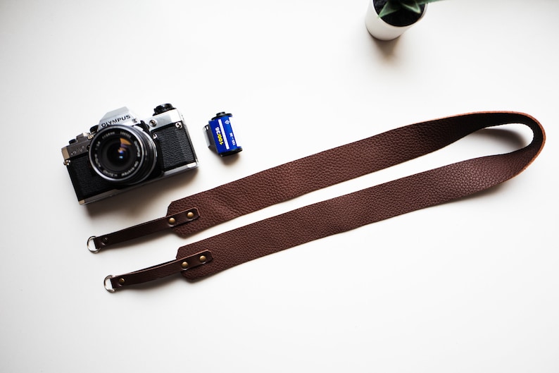 May include: A brown leather camera strap with metal hardware and two adjustable loops. The strap is laid out flat on a white surface.