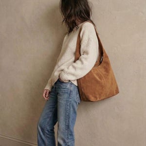 May include: A tan suede shoulder bag with a zipper closure is worn over a cream-colored sweater and blue jeans. The bag is a medium size and has a simple, casual design. The outfit is leaning against a neutral-colored wall.