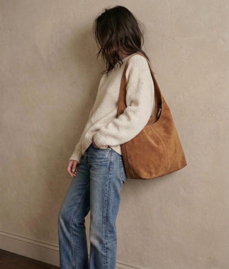 May include: A tan suede shoulder bag with a zipper closure is worn over a cream-colored sweater and blue jeans. The bag is a medium size and has a simple, casual design. The outfit is leaning against a neutral-colored wall.