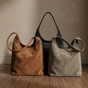 May include: Three suede shoulder bags in shades of brown, gray, and black. The bags have a soft, textured appearance and feature a single shoulder strap. The bags are displayed on a wooden floor, showcasing their shape and design.