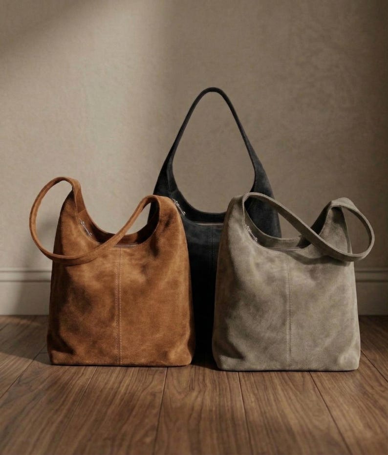 May include: Three suede shoulder bags in shades of brown, gray, and black. The bags have a soft, textured appearance and feature a single shoulder strap. The bags are displayed on a wooden floor, showcasing their shape and design.