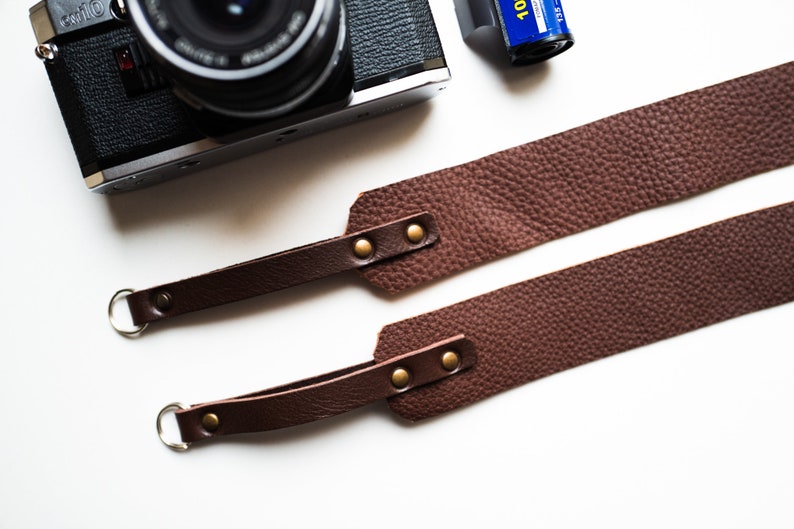 May include: A brown leather camera strap with two metal rings and gold-coloured rivets. The strap is lying flat on a white surface.