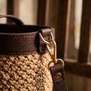 May include: A bucket bag featuring a woven tan straw body and a dark brown pebbled leather top trim. The bag includes a dark brown leather handle and a detachable dark brown leather shoulder strap connected by polished gold-tone metal hardware.