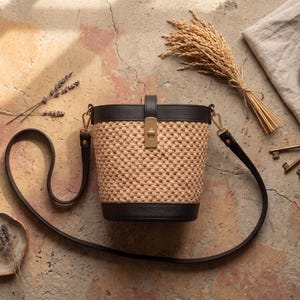 May include: A bucket bag featuring a woven tan straw body with dark brown leather trim at the top and bottom. The bag includes a gold-toned metal turn-lock closure on the front and a long, adjustable dark brown leather shoulder strap.