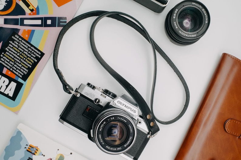 May include: A silver and black 35mm film camera with a black leather strap. The camera has a silver lens with a black lens cap. The camera is sitting on a white surface.