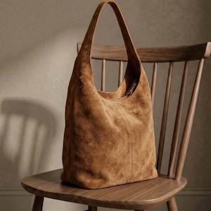 May include: A large, brown suede tote bag with a single shoulder strap. The bag is sitting on a wooden chair with a curved back. The suede has a soft, textured appearance, and the bag appears to be empty.