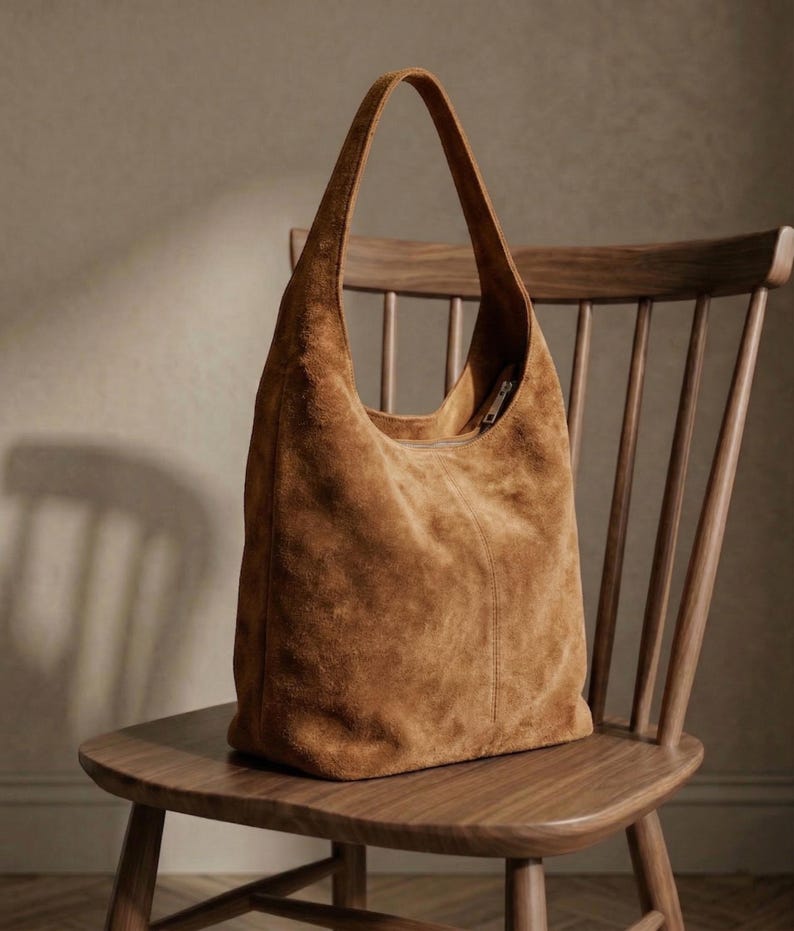 May include: A large, brown suede tote bag with a single shoulder strap. The bag is sitting on a wooden chair with a curved back. The suede has a soft, textured appearance, and the bag appears to be empty.