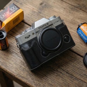 Könnte beinhalten: Eine silberne Fujifilm X-T30 Kamera mit schwarzer Ledertasche und Objektivdeckel, neben Filmrollen Ektar 100 und Cinestill 800T. Die Kamera und der Film liegen auf einer Holzoberfläche, was ein Vintage-Fotografie-Thema erzeugt.