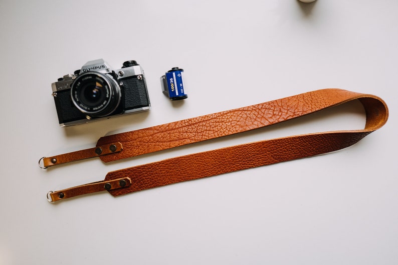 May include: A brown leather camera strap with metal rings and two adjustable straps. The strap is lying on a white surface next to a silver camera and a roll of film.