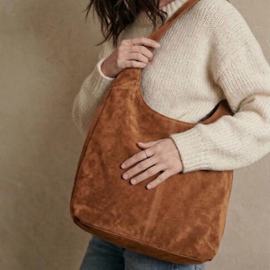 May include: A brown suede shoulder bag with a curved design. The bag has a single strap and is held by a person wearing a cream-colored sweater and blue jeans. The bag's texture is visible.