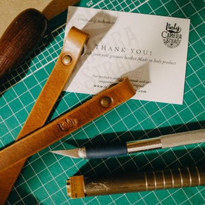 May include: A brown leather camera strap with the words "Holy Camera Straps" stamped on it. The strap is lying on a green cutting mat with a thank you card and various tools.