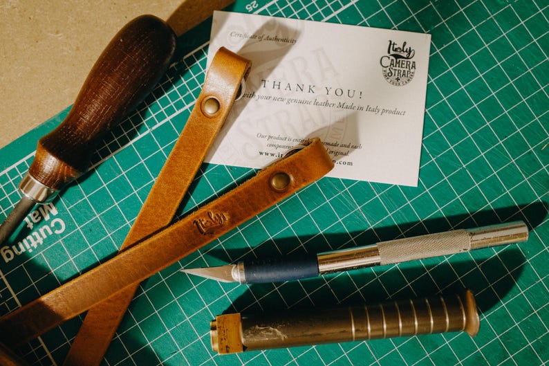 May include: A brown leather camera strap with the words "Holy Camera Straps" stamped on it. The strap is lying on a green cutting mat with a thank you card and various tools.