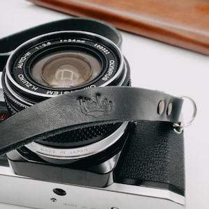 May include: A black leather camera strap with a silver metal ring and the word "Hello" embossed on the strap. The strap is attached to a black camera lens with silver accents. The lens has the text "AUTO-M  1:2.8  f=24mm  100721  OLYMPUS  ZUIKO  H.ZUIKO  SYSTEM  MADE IN JAPAN" printed on it.
