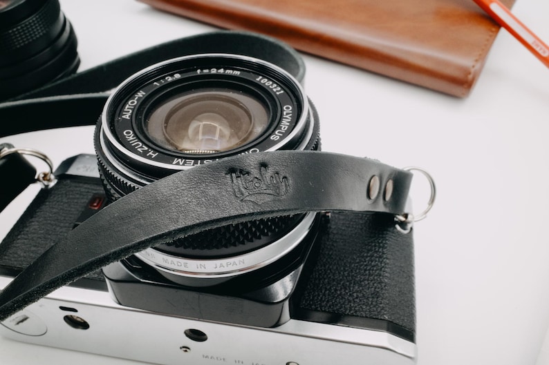 May include: A black leather camera strap with a silver metal ring and the word "Hello" embossed on the strap. The strap is attached to a black camera lens with silver accents. The lens has the text "AUTO-M  1:2.8  f=24mm  100721  OLYMPUS  ZUIKO  H.ZUIKO  SYSTEM  MADE IN JAPAN" printed on it.