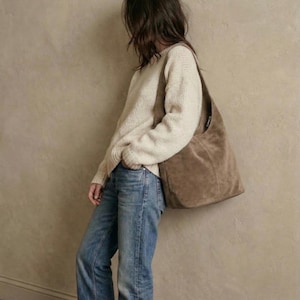 May include: A person wearing a cream-colored sweater, blue jeans, and a large brown suede shoulder bag. The jeans have a frayed hem. The bag is large and unstructured, with a single strap.