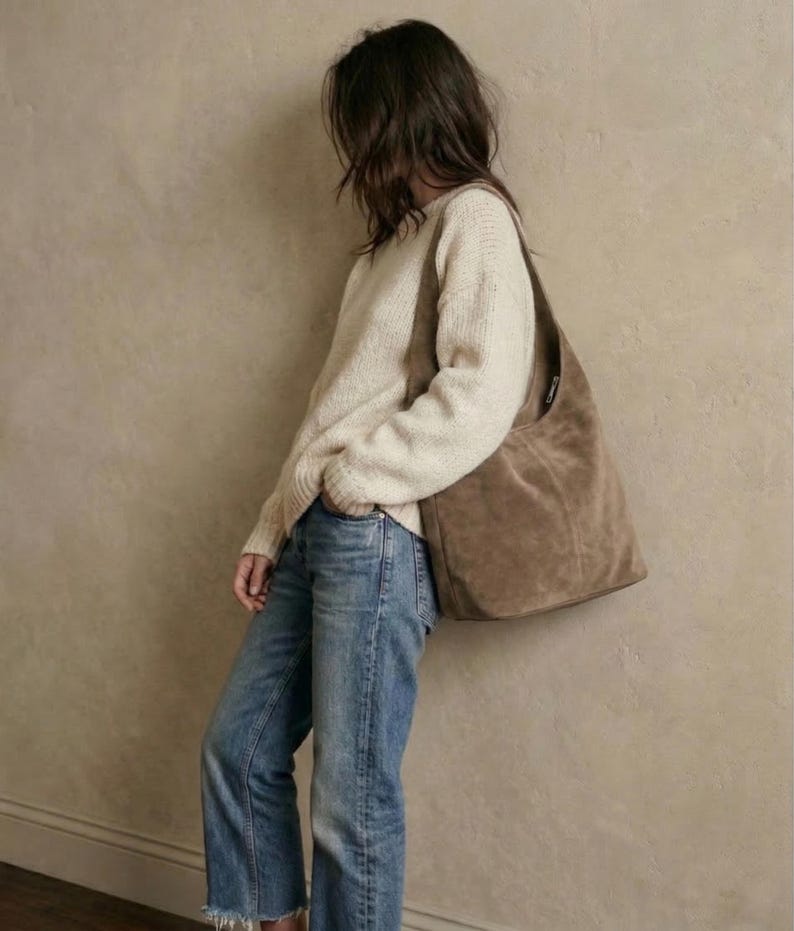 May include: A person wearing a cream-colored sweater, blue jeans, and a large brown suede shoulder bag. The jeans have a frayed hem. The bag is large and unstructured, with a single strap.