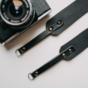 May include: Black leather camera strap with two metal rings and gold-toned rivets. The strap is laid out flat on a white surface.