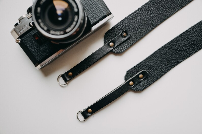 May include: Black leather camera strap with two metal rings and gold-toned rivets. The strap is laid out flat on a white surface.