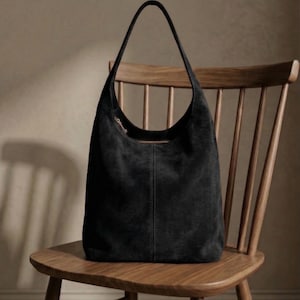 May include: A black suede shoulder bag with a curved top and long strap, resting on a wooden chair. The bag has a zipper closure and a vertical seam down the center. The chair is made of light-colored wood.