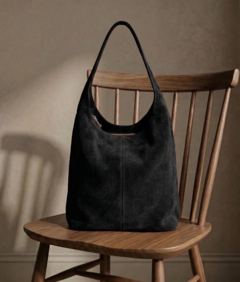 May include: A black suede shoulder bag with a curved top and long strap, resting on a wooden chair. The bag has a zipper closure and a vertical seam down the center. The chair is made of light-colored wood.