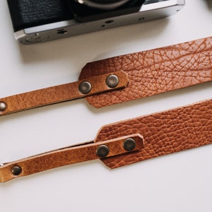 May include: Two brown leather camera straps with metal rings and snaps. The straps are made of textured leather and have a rustic look.