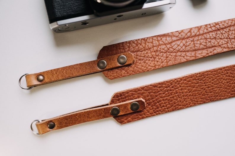 May include: Two brown leather camera straps with metal rings and snaps. The straps are made of textured leather and have a rustic look.