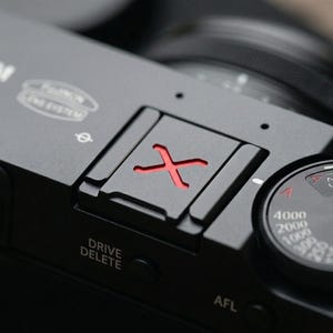 Puede incluir: Primer plano de un cuerpo de cámara negro con una marca 'X' roja en la parte superior. La cámara tiene un dial con números y un botón con la etiqueta 'DRIVE DELETE'. El logotipo de Fujifilm es visible.