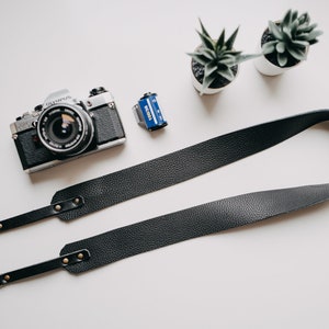 May include: A black leather camera strap with gold metal hardware and two D-rings. The strap is lying on a white surface next to a silver camera and a film canister.