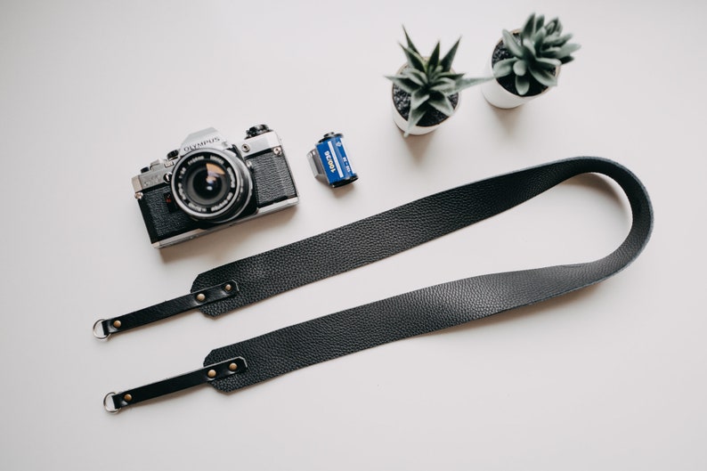 May include: A black leather camera strap with gold metal hardware and two D-rings. The strap is lying on a white surface next to a silver camera and a film canister.