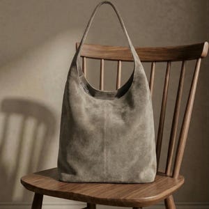May include: A taupe suede hobo bag with a single shoulder strap. The bag has a curved top and a vertical seam down the center. It rests on a wooden chair with a curved back and spindle supports. The bag's soft texture contrasts with the chair's smooth wood.