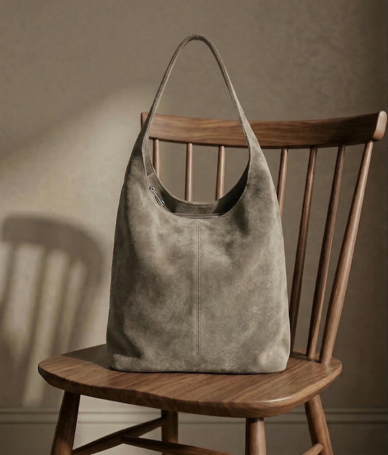 May include: A taupe suede hobo bag with a single shoulder strap. The bag has a curved top and a vertical seam down the center. It rests on a wooden chair with a curved back and spindle supports. The bag's soft texture contrasts with the chair's smooth wood.