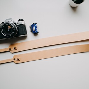 May include: A tan leather camera strap with two metal rings and brass rivets. The strap is folded in half and laid flat on a white surface. A silver film camera and a blue film canister are also visible in the image.