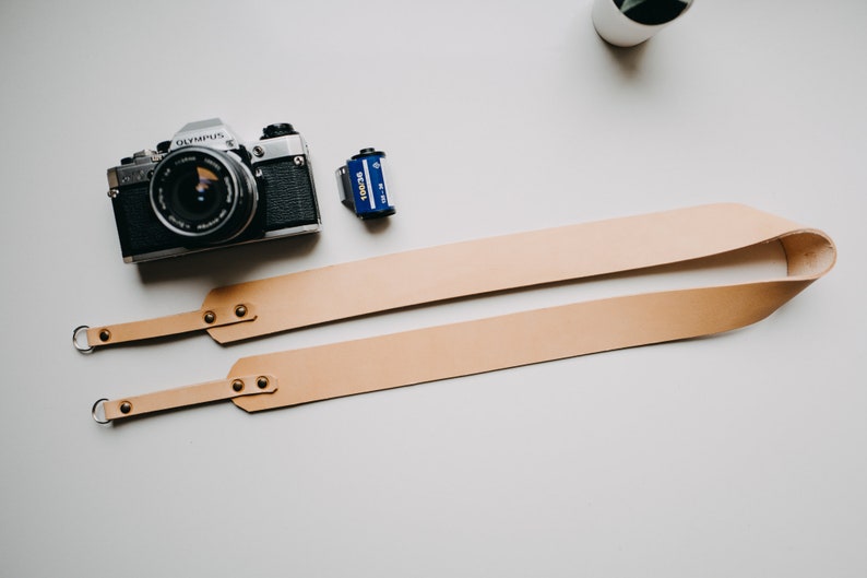 May include: A tan leather camera strap with two metal rings and brass rivets. The strap is folded in half and laid flat on a white surface. A silver film camera and a blue film canister are also visible in the image.