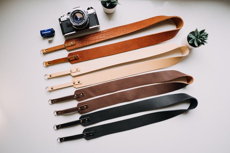 May include: Five different leather camera straps in various colours, including brown, tan, dark brown, and black. The straps are laid out in a row on a white surface.