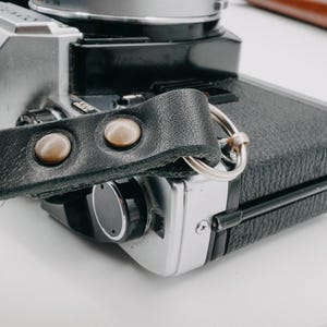 May include: A black leather camera strap with two silver metal accents. The strap is attached to a silver and black camera.