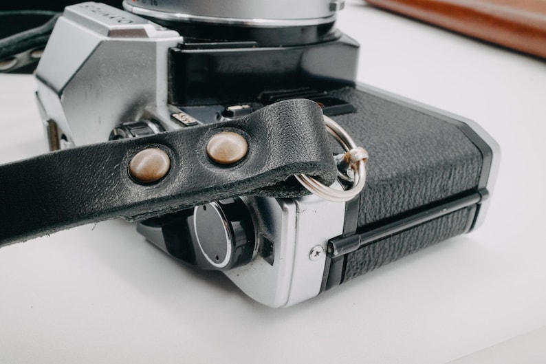 May include: A black leather camera strap with two silver metal accents. The strap is attached to a silver and black camera.