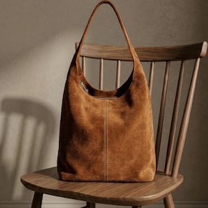 Puede incluir: Un bolso de hombro de ante marrón con una correa larga, apoyado en una silla de madera. El bolso tiene una parte superior curva y una costura vertical en el centro. La silla tiene respaldo de listones y asiento curvo.
