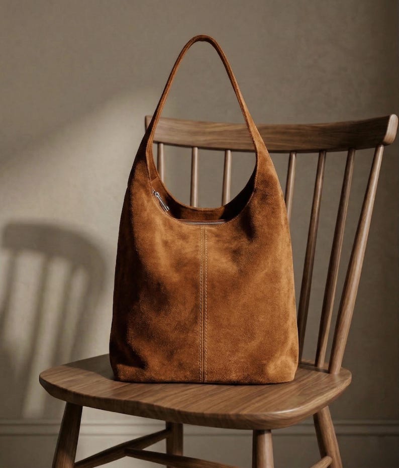May include: A brown suede shoulder bag with a long strap, resting on a wooden chair. The bag has a curved top and a vertical seam down the center. The chair has a slatted back and a curved seat.