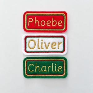 May include: Three rectangular embroidered name patches. The top patch is red with gold text reading Phoebe. The middle patch is white with gold text reading Oliver. The bottom patch is green with gold text reading Charlie. Each has a border.