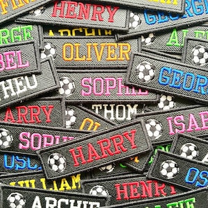 May include: A selection of black name patches with soccer ball designs. Names such as "HENRY", "OLIVER", and "HARRY" are embroidered in colours like red, blue, and yellow. The patches are rectangular in shape.
