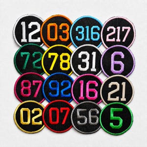 May include: A collection of sixteen circular embroidered patches arranged in a four-by-four grid. Each patch features a black background with a colored border and a large, bold number stitched in the center in a matching or contrasting thread color.