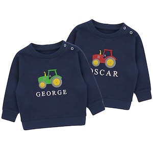 May include: Two navy blue children's sweatshirts. One has a green tractor and the name "GEORGE" embroidered in white. The other has a red tractor and the name "OSCAR" embroidered in white. Both sweatshirts have yellow wheels and small shoulder snaps.