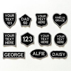May include: A collection of black and white embroidered patches in various shapes, including shields, hearts, and rectangles. Each patch features white text, with some displaying names like "GEORGE" and "DAISY", and others customizable with "YOUR TEXT HERE".